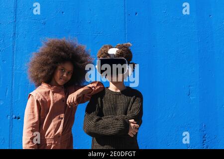 Ragazza nera teen positiva con capelli ricci in piedi vicino irriconoscibile Un amico indossa il visore VR mentre passa il tempo insieme e sperimenta virtuale Foto Stock