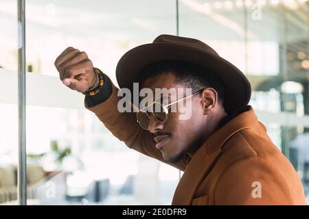 Self esteem giovane ragazzo etnico sopportato in occhiali da sole alla moda e. cappello appoggiato su una parete di vetro di un edificio moderno Foto Stock