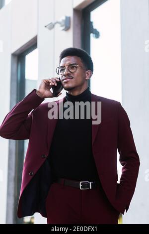 Serio giovane Africano americano manager maschile in abito elegante e. occhiali che parlano di problemi aziendali sullo smartphone e si guardano via Foto Stock