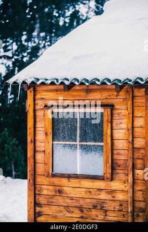 Accogliente tipica casa in legno con tetto innevato e piccola finestra coperto di brina il giorno d'inverno Foto Stock