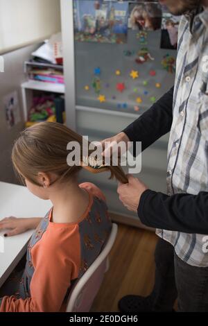 Vista laterale di un uomo adulto positivo tagliato e non riconoscibile che spazzolava a lungo capelli di figlia preseen con spazzola durante la routine quotidiana a. casa Foto Stock