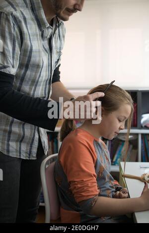 Vista laterale di un uomo adulto positivo tagliato e non riconoscibile che spazzolava a lungo capelli di figlia preseen con spazzola durante la routine quotidiana a. casa Foto Stock