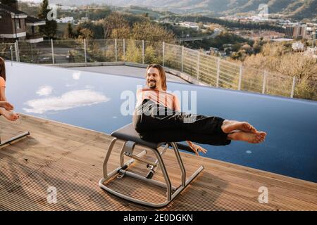 Felice maschio in activewear equilibrare su pilates sedia e fare abs esercizi mentre sorridendo durante l'allenamento in terrazza e guardando via Foto Stock