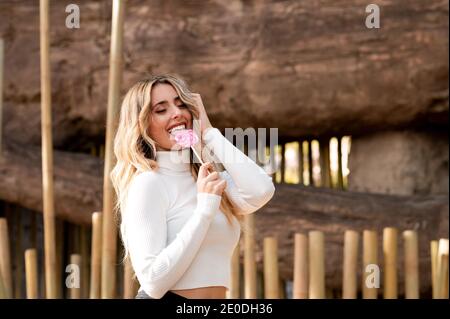 Vista laterale di giovane femmina di cotetta in piedi nel parco e. leccare il dolce lecca rosa mentre godete il fine settimana con gli occhi chiusi Foto Stock