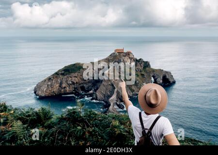 Vista posteriore del turista maschile irriconoscibile in piedi dall'alto osservando Scenario dell'isola di Gaztelugatxe in mare durante le vacanze estive Foto Stock