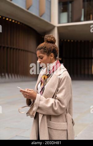 Vista laterale della giovane imprenditrice afroamericana con riccia capelli in elegante cappotto utilizzando smartphone durante l'attraversamento della strada Londra con laptop in Foto Stock