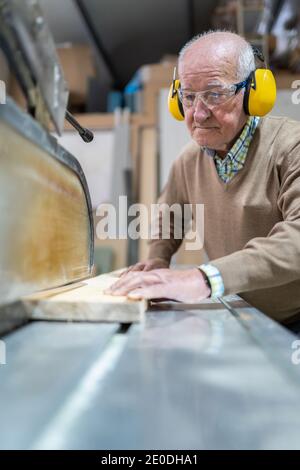 Uomo anziano adulto concentrato artigiano in occhiali e cuffie segatura tavola di legno con sega circolare affilata in officina Foto Stock