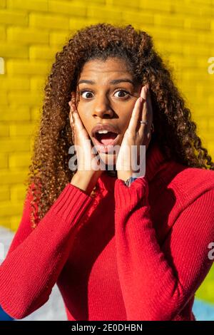 Sorprendente femmina afroamericana adolescente con lunghi capelli ricci dentro pullover rosso in piedi vicino a colorati muri di graffiti con bocca aperta e mani su ch Foto Stock