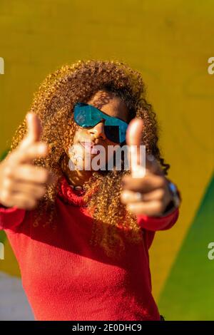 Giovane donna afroamericana allegra con capelli ricci casual vestiti e occhiali da sole sorridenti e puntano la fotocamera contro la luce parete colorata Foto Stock