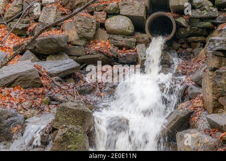 Un torrente di acqua che sgorga da un tubo di cemento in alto sul lato di una collina rocciosa in una giornata piovosa. Foglie morte e rami a terra. Foto Stock