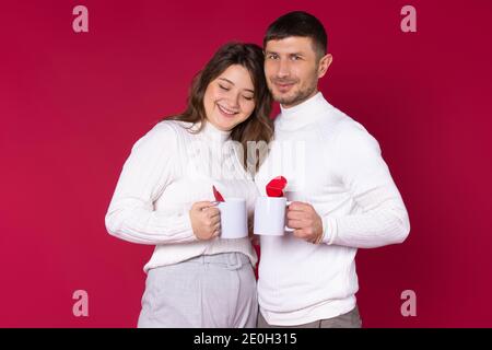 Una coppia amorevole con due tazze bianche di tè per il vostro aggiungere sta sorridendo su uno sfondo rosso. Cuori rossi . Foto di alta qualità Foto Stock