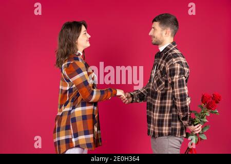 Giovane coppia innamorato in studio su sfondo rosso, tenere le mani. L'uomo nasconde le rose dietro la schiena. Foto di alta qualità Foto Stock