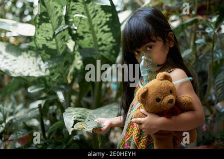 Carina bambina con maschera di inalazione nebulizzatore che la tiene Tedy Orso nel parco Foto Stock