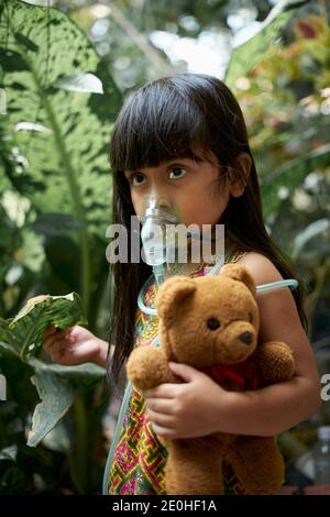 Carina bambina con maschera di inalazione nebulizzatore che la tiene Tedy Orso nel parco Foto Stock