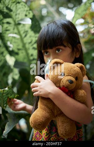 Carina bambina con maschera di inalazione nebulizzatore che la tiene Tedy Orso nel parco Foto Stock