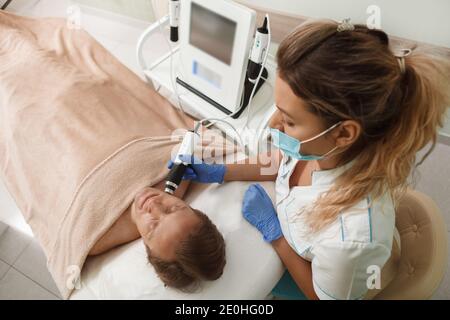 Vista dall'alto di un cosmetologo professionista che indossa il viso medico maschera mentre si lavora con il client maschile Foto Stock