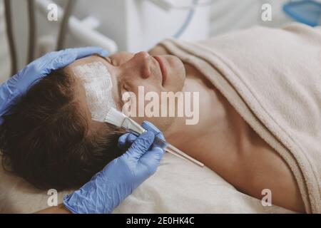 Primo piano di un bell'uomo maturo rilassato che ottiene il trattamento della maschera facciale al centro termale. Rilassante cliente maschile felice godendo trattamento skincare a cosmesi Foto Stock