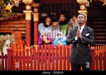 Ritratto di giovane e bell'uomo d'affari afro-americano in tuta soggiorno e pregare contro presepe di Natale e scena presepe. Foto Stock