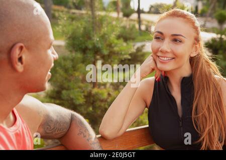 Bella donna felice sorridendo gioiosamente al suo ragazzo, riposandosi insieme all'aperto dopo essersi allenati. Una coppia affascinante che parla, seduto sulla panchina Foto Stock
