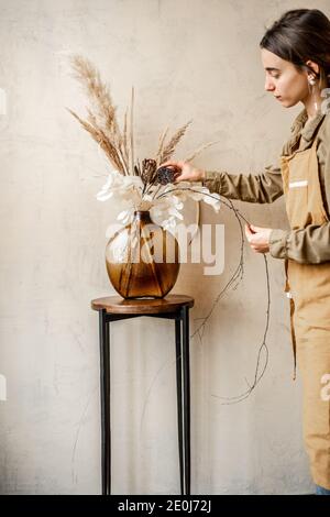 Donna decorazione casa con una composizione di fiori secchi e. erbe in un vaso di vetro su uno sfondo beige Foto Stock