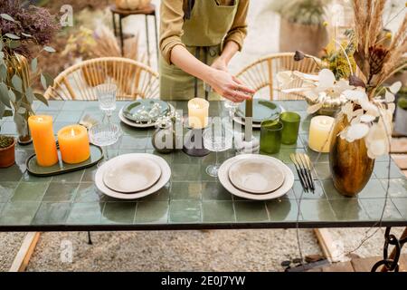 Giovane donna che illumina le candele mentre decorando la tavola da pranzo in naturale Stile boho in toni verdi all'aperto Foto Stock