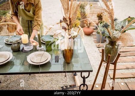 Giovane donna che illumina le candele mentre decorando la tavola da pranzo in naturale Stile boho in toni verdi all'aperto Foto Stock