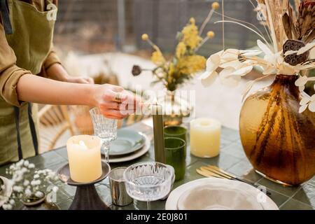 Giovane donna che illumina le candele mentre decorando la tavola da pranzo in naturale Stile boho in toni verdi all'aperto Foto Stock