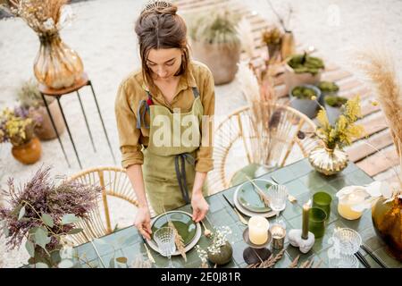 Giovane decoratore femminile o fiorista che mette le erbe sul tavolo, decorando il pranzo posto in stile naturale Boho in toni verdi all'aperto Foto Stock