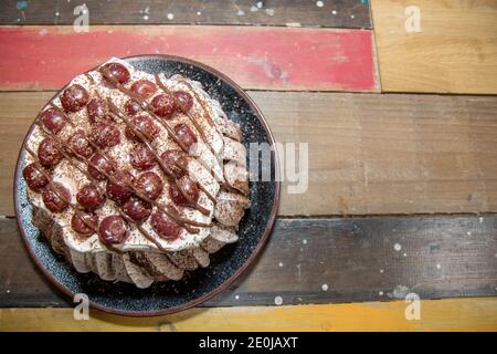 Un delizioso deserto di pavlova con crema, ciliegie e cioccolato sciroppo su un piatto marrone su un lavoro di cucina di legno in alto Foto Stock