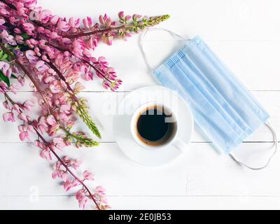 Tazza di caffè, maschera medica rimossa e fiori rosa. Colazione nel caffè. Realtà moderna e concetto di vita. Maschera chirurgica protettiva blu e bianca Foto Stock