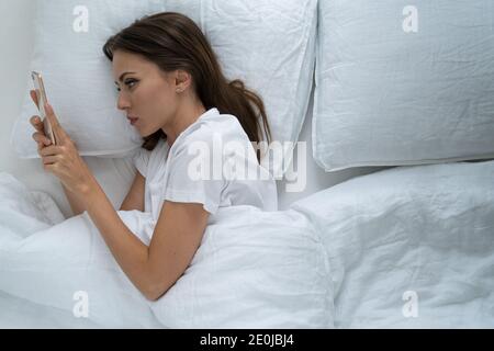 Donna solitaria con espressione del viso depressa che si trova a letto tenendo mobile smartphone, non può dormire, dipendente dai social network, guardando lo schermo e. Foto Stock
