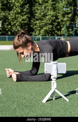 Donna in sportswear vlogger seduta su tappeto erboso artificiale all'aperto, mostrando e dimostrando esercizi per il suo blog online, record su fotocamera smartphone Foto Stock