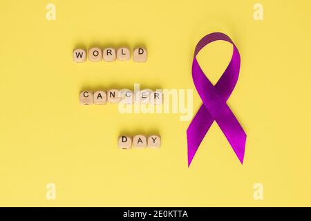 Cubi di legno con testo della Giornata Mondiale del cancro e nastro viola, su sfondo giallo. Disposizione piatta Foto Stock