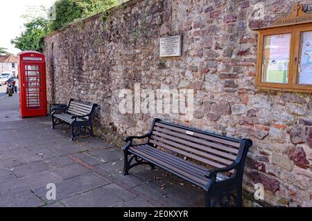 Due panchine pubbliche in legno e una scatola telefonica si trovano nelle loro parti Un muro di pietra naturale nel centro del villaggio di Nether Stowey nel Somerset Foto Stock