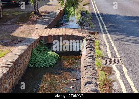 Un piccolo ruscello scorre attraverso gli alberi e sotto un piccolo ponte in un idilliaco villaggio Somerset. Foto Stock