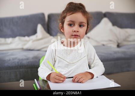 Una bella ragazza bebè curiosa seduta ad un tavolo tiene una penna e guarda via. Disegno a casa. Foto Stock