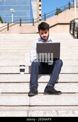 Giovane uomo d'affari che usa il suo computer portatile, con una tazza di caffè, seduto sulle scale. Uomo d'affari in una strada urbana di una città. Foto Stock