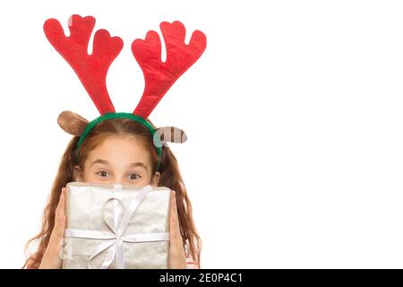 Un colpo corto di una ragazza felice piccola che indossa Natale della renna antlers che nascondono il suo volto dietro una scatola di regalo che guarda a. la telecamera da oltre un iso presente Foto Stock