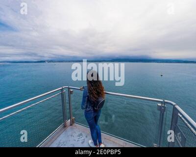 Giovane bruna vestita in denim in un gazebo di vetro di fronte al mare. Messa a fuoco selezionata. Foto Stock