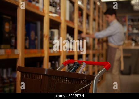 Fuoco selettivo su un carrello della spesa con le bottiglie, il proprietario del negozio del vino che organizza le bevande sugli scaffali sullo sfondo Foto Stock
