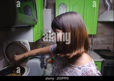 sorridente giovane donna dai capelli neri mescolando riso con verdure usando una spatola del raschietto della cucina, che è preparata per la cena in un multicooker nella k Foto Stock