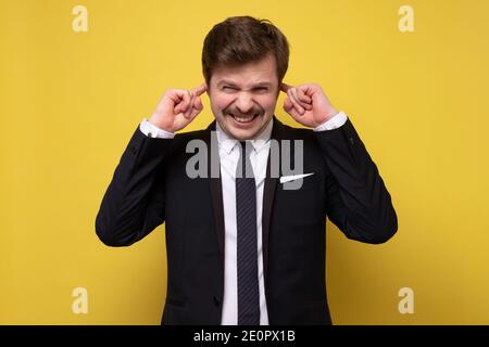 Questo è troppo forte. Frustrato giovane uomo in formalwear tappando le orecchie con le dita e mantenendo gli occhi chiusi. Studio girato su parete gialla. Foto Stock