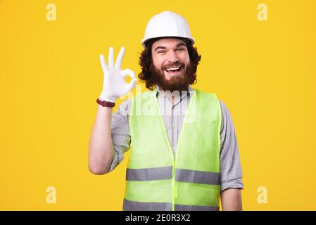 Un bel giovane architetto bearded sta guardando allegro alla telecamera che mostra il segno ok vicino a una parete gialla . Foto Stock