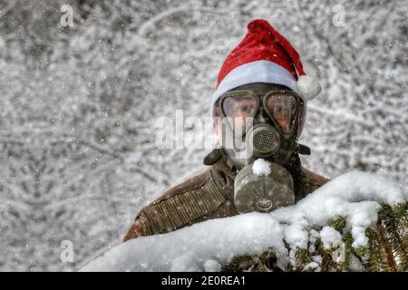 Dietro un ramo innevato di abete si trova un uomo con una maschera a gas e un cappello rosso Santa in una nevicata nel paesaggio invernale. Foto Stock