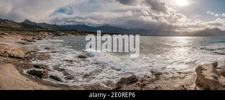 Vista panoramica delle onde che si lavano sulla costa rocciosa di Calvi Bay nella regione Balagne della Corsica con neve montagne ricoperte in lontananza Foto Stock