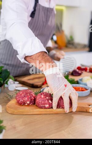 Chef che prepara polpette e le mette su una tavola di legno. Ci sono varie spezie, verdure, così come piccole bocce da cucina intorno al lavoro surf Foto Stock