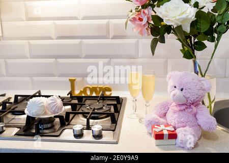 Adorabile orsacchiotto di bambola con rose rosa bianche, piccola scatola regalo, marshmallow e champagne in cucina vicino alla stufa per il concetto di San Valentino Foto Stock