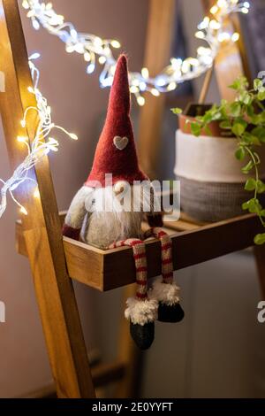 Elfo di Natale seduto su una mensola di legno accanto ad una pianta verde, illuminata da ghirlande leggere. Carino decorazione di natale. Foto Stock