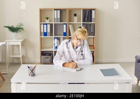 Serio medico maturo seduto alla scrivania, lavorando con i documenti e scrivendo le storie mediche Foto Stock