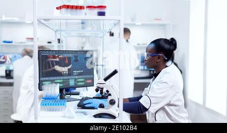 Scienziato africano che lavora al computer in una struttura moderna e colleghi in background. Nero reseacher sanitario in laboratorio di biochimica indossando attrezzature sterili. Foto Stock